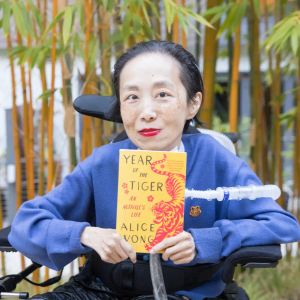 Photo of Alice Wong, an Asian American disabled woman in a power chair, against a background of bamboo trees. She is wearing a blue cardigan and sitting in a power chair. She is holding a copy of her memoir, Year of the Tiger, a paperback in yellow and red with a fierce tiger on it designed by Madeline Partner. She is wearing a bold red lip color and a trach at her neck.