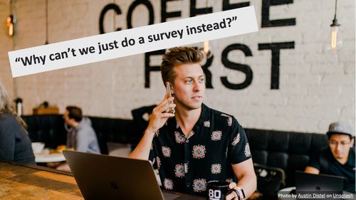 A man on the phone, sat at a computer, in a coffee shop. The overlaid text reads "Why can't we just do a survey instead". There is a photo credit to Austin Distel on Unsplash.